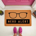 Nerd Alert doormat featuring black glasses design on a natural coir welcome mat, placed on a porch with books and a plant, making a funny front door rug for home decor, entryway floor mat, or a unique housewarming gift for book