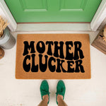 Funny Mother Clucker doormat featuring bold black text on a natural coir fiber welcome mat, placed on a front porch in front of a green door with a person wearing green sneakers standing on the mat for a humorous home entryway