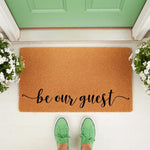 Be Our Guest coir doormat featuring natural fiber construction with black script lettering, placed on a front porch entryway with green sneakers visible, making a stylish welcome mat for home decor and housewarming entryways.