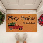 Personalized Merry Christmas doormat featuring a red truck carrying a pine tree, this custom family name welcome mat is a festive holiday porch decor piece for your home entrance, shown on a front porch with a plaid blanket and
