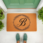 Personalized monogram doormat featuring a black letter B inside a circular leaf wreath design on natural coir fiber, perfect as a custom front porch welcome mat for a housewarming entry or a stylish home decor door rug.