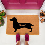 Personalized Dachshund doormat featuring a black dog silhouette on natural coir fiber, perfect as a custom pet lover welcome mat for your front porch entry, shown on a white floor in front of a bright pink door with potted plants.