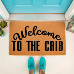 Welcome to the Crib doormat featuring bold black text on a natural coir fiber mat, perfect for a funny front porch entrance or housewarming decor, shown on a white floor in front of a bright blue door with a person wearing teal