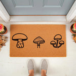 Mushroom doormat with a natural coir fiber texture, featuring three black line art fungi illustrations for a cottagecore porch decor look, placed on a front entryway floor with potted plants and a person wearing gray slip-on