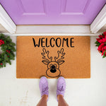 Reindeer welcome doormat for Christmas porch decor, featuring a natural coir fiber mat with a black printed reindeer face and welcome text, placed on a front porch with festive greenery and a purple door for a holiday entryway