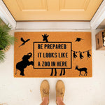 Zoo animal welcome doormat featuring a funny quote about a zoo in here, with black silhouettes of an elephant, giraffe, monkeys, and birds on a natural coir fiber mat placed at a front door entrance for a fun home decor entryway.