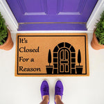 It's Closed For a Reason doormat, a funny natural coir welcome mat with a black door graphic, placed on a porch in front of a purple door with potted plants, perfect for a humorous home entrance or a quirky front door decor