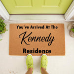 Personalized family name doormat featuring the text You've Arrived At The Residence on a natural coir fiber welcome mat, placed on a front porch with lime green sneakers visible, creating a stylish custom housewarming entry decor.