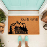 Cabin Fever mountain house doormat featuring a rustic log cabin and pine tree design on natural coir fiber, perfect for a front porch welcome mat or outdoor entryway decor for nature lovers and mountain home enthusiasts.