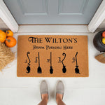 Personalized family broom parking doormat for Halloween, featuring a natural coir welcome mat with custom names and witch broom illustrations, perfect for a festive fall porch decor setup with pumpkins and a black cauldron nearby.