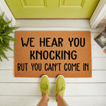 Funny doormat with the text We Hear You Knocking But You Can't Come In, a natural coir fiber welcome mat for front porch decor that adds a humorous touch to your home entrance and entryway floor with its bold black lettering