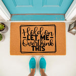 Funny Hold On Let Me Overthink This doormat featuring bold black text on a natural coir fiber welcome mat, placed on a porch in front of a bright blue door with a person wearing teal slip-on shoes standing on the front door mat.