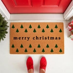 Merry Christmas doormat with green pine tree pattern on natural coir fiber, featuring a festive holiday welcome mat design for front porch decor, shown on a white floor with red shoes and a bright red door in the background.