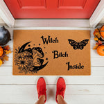 Witch Bitch Inside coir doormat featuring a crescent moon, mushroom, and butterfly design for a funny front porch welcome mat, perfect for fall home decor or a spooky entryway rug placed on a white floor by a red door.
