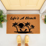 Life's A Beach tropical doormat featuring a sunset, palm trees, and lounge chairs on a natural coir welcome mat, perfect for a coastal home entrance, summer porch decor, or a beach house front door rug for outdoor entryways.