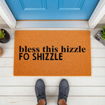 Funny doormat with the text bless this hizzle fo shizzle printed in black on a natural coir welcome mat, placed on a white porch in front of a blue door for a humorous home entrance decor look that greets guests with personality.