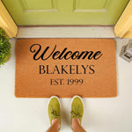 Personalized family name welcome doormat featuring custom text and established year on a natural coir fiber mat, placed on a front porch entryway with a person wearing green sneakers standing on the durable outdoor floor rug.