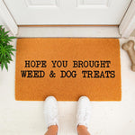 Funny weed and dog treats doormat featuring bold black text on a natural coir fiber welcome mat, perfect for a porch entry way with a plant and dog bone toy nearby, making this a humorous housewarming decor piece for pet owners.