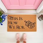 Hanukkah coir doormat featuring a dreidel design and the text This Is How We Roll, a natural fiber welcome mat for holiday home decor placed on a porch in front of a pink door with a person wearing pink sneakers standing nearby.