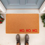 Ho Ho Ho Christmas doormat featuring natural coir fiber material with bold red text, placed on a front porch entryway with festive holiday greenery decor and a person standing on the welcome mat in casual gray shoes.