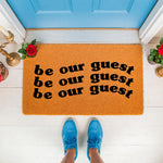 Be our guest doormat featuring a natural fiber coir welcome mat with black text, placed on a white porch in front of a blue door, perfect for a front entryway, housewarming decor, or a stylish outdoor floor mat for your home
