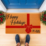 Personalized Happy Holidays doormat featuring a red gift bow design on a natural coir welcome mat, perfect for Christmas porch decor with custom family name text, shown on a front porch with festive greenery and a blue door