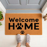 Pet lover welcome home doormat with black paw print design on a natural fiber mat, placed at a front door entrance for dog owners who want a cute entryway rug or porch decor that celebrates their furry friend and home entrance.