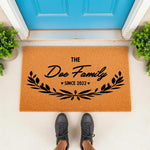 Personalized family name doormat featuring a natural coir fiber texture with black text and a botanical leaf design, perfect as a custom housewarming welcome mat for a front porch entrance to celebrate a new home or wedding