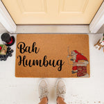 Bah Humbug Christmas doormat featuring a funny illustration of a man in a red suit sitting on a chimney, this natural coir welcome mat is a humorous holiday floor rug for your front porch decor during the winter festive season.