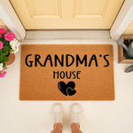Grandma's House coir doormat with black text and heart icons, placed on a porch in front of a door with flowers and a watering can, making a stylish welcome mat for a home entrance or a personalized housewarming decor item.