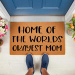 Funny Mom Doormat with Home of the Worlds Okayest Mom text on a natural coir welcome mat, placed on a porch in front of a blue door, making a humorous housewarming gift for a mom who appreciates funny home decor and entryway rugs.
