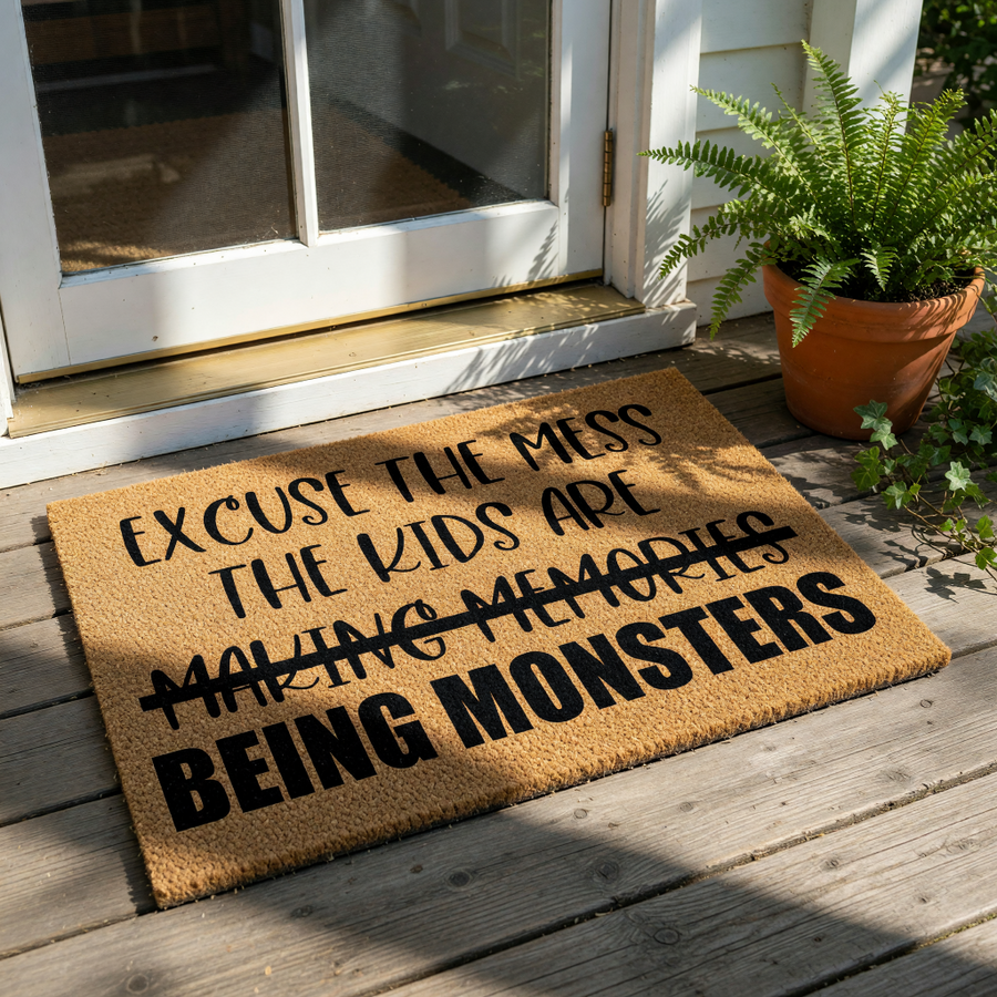 Funny coir doormat with the text Excuse the mess the kids are making memories being monsters, a humorous welcome mat for parents that features a crossed-out phrase and bold black lettering on a natural brown fiber floor rug.