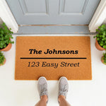 Personalized family name doormat featuring a custom address, this natural coir fiber welcome mat sits on a porch with potted plants, providing a stylish housewarming entrance decor for your home front door and entryway floor.