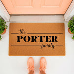 Personalized family name coir doormat for front porch entry, featuring a natural fiber welcome mat with custom text design, placed on a white floor in front of a peach door with potted plants and a person wearing peach sneakers.