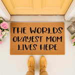 Funny Mom Doormat with the text The Worlds Okayest Mom Lives Here, featuring a natural coir fiber welcome mat on a front porch, making a humorous home decor entry rug for a mom who appreciates funny housewarming or birthday gifts.