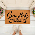 Grandkids make life grand doormat for grandparents, a natural coir welcome mat with black script text and heart accents placed on a white porch floor with potted flowers and toys for a cute entryway decor look.