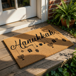 Hanukkah coir doormat featuring a natural fiber texture with black script text and hanging holiday icons like menorahs, stars, and gift boxes, making a festive welcome mat for your front porch during the winter holiday season.