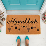 Hanukkah doormat with festive holiday icons like menorahs, dreidels, and stars hanging from the text, this natural coir welcome mat creates a stylish entryway decor piece for the winter season on a front porch with teal door.