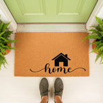 Home coir doormat with a black house icon and cursive script text, featuring a natural fiber welcome mat design for a front porch entry, placed on a white floor in front of a green door with potted ferns for a stylish