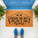 Pardon Our Mess But This Is Our Home doormat featuring a house outline and heart design on natural coir fiber, perfect as a funny front porch welcome mat for a housewarming or new home decor, shown on a white porch with blue door.