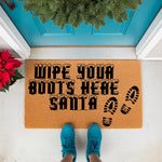 Wipe Your Boots Here Santa Christmas doormat featuring black text with snow accents and boot prints on a natural coir fiber welcome mat, placed on a front porch for holiday home decor and festive winter entryway floor mat styling.