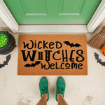 Wicked Witches Welcome Halloween doormat featuring black bat graphics on a natural coir fiber mat, perfect for a spooky front porch entryway decor, placed on a concrete step in front of a green door with a person wearing green