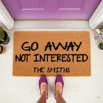 Personalized funny doormat with Go Away Not Interested text on a natural coir welcome mat, featuring a custom family name sign for your front porch decor, shown in a top-down view with purple shoes standing on the outdoor floor