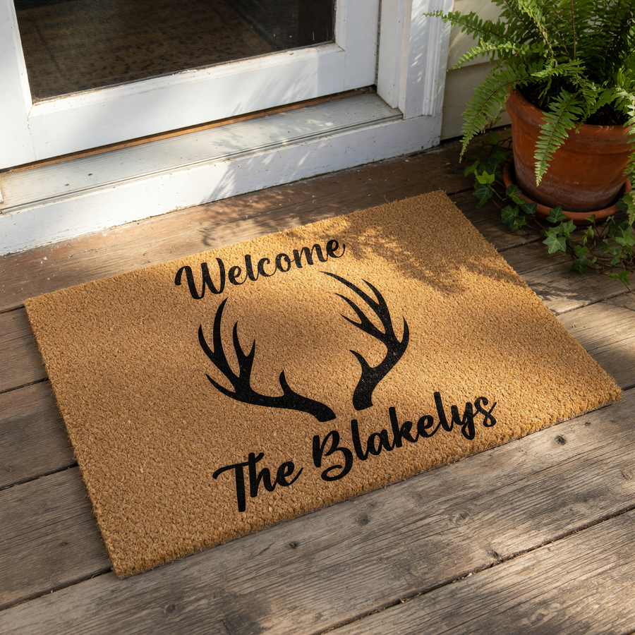 Personalized antler welcome doormat with rustic outdoor decor style, featuring a natural coir fiber texture and black printed text for a custom family name entry rug that makes a stylish housewarming front porch floor mat for any