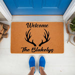 Personalized antler welcome doormat featuring a rustic deer horn design and custom family name text, displayed on a front porch entrance mat for home decor, perfect for a cabin or lodge style entryway with a natural fiber floor