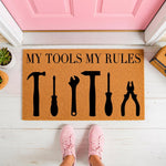 My Tools My Rules doormat featuring black tool silhouettes on a natural coir welcome mat, perfect for a garage or workshop entrance, shown on a porch floor with pink sneakers standing in front of the funny handyman home decor.
