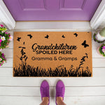 Gramma and Gramps doormat featuring a playful grandchildren spoiled here design with butterflies and grass, this natural coir welcome mat is a fun outdoor porch decor piece for a grandparent home entrance on a white deck.