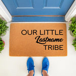 Personalized family name doormat featuring the text Our Little Lastname Tribe on a natural coir fiber welcome mat, placed on a front porch entryway with blue shoes visible, perfect for a custom housewarming or new home decor