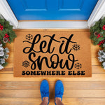 Let It Snow Somewhere Else funny winter doormat featuring black text and snowflakes on a natural coir fiber welcome mat, placed on a front porch with festive greenery and blue shoes visible in this holiday home decor scene.