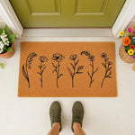 Floral line art doormat featuring a natural coir welcome mat with black botanical wildflower illustrations, placed on a porch in front of a green door with potted plants and a person wearing olive green sneakers standing on the